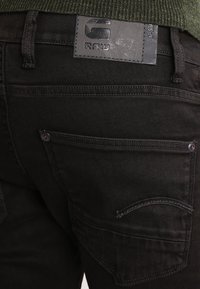Jeans de mezclilla negra con una etiqueta de cuero texturizado en la pretina. Los bolsillos traseros tienen bordes redondeados con detalles de costura en contraste.