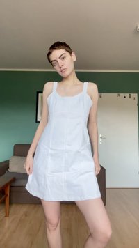 Νεαρό άτομο με κοντά μαλλιά στέκεται σε ένα δωμάτιο φορώντας ένα λευκό φόρεμα χωρίς μανίκια, με έναν καναπέ και μια κλειστή πόρτα στο φόντο.