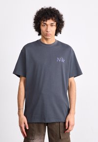 T-shirt de algodão cinza com mangas curtas, apresentando um pequeno logótipo roxo no peito. Corte solto e decote redondo.