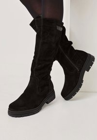 Knee-high black suede boots featuring a textured finish, side zipper, and a rugged black rubber sole with defined tread.