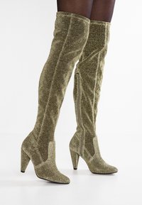 Stivali sopra il ginocchio verde oliva lucidi in un tessuto testurizzato. Presentano tacco a stiletto alto e una cerniera laterale per la chiusura.