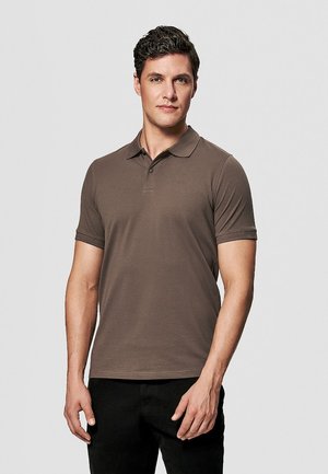 Mężczyzna stojący o neutralnym wyrazie twarzy, ubrany w brązową koszulkę polo z krótkim rękawem i czarne spodnie na jednolitym, jasnym tle.