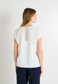 Haut blanc à manches courtes avec un détail en dentelle au niveau de l'encolure arrière. Tissu doux avec une coupe décontractée, associé à un pantalon bleu marine.