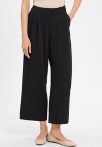 Black wide-leg trousers with a textured elastic waistband, featuring front pleats and a smooth fabric. Paired with light-colored flats.