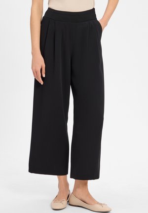Schwarze weit geschnittene Hose mit einem strukturierten Gummibund, ausgestattet mit vorderen Falten und einem glatten Stoff. Kombiniert mit hellen Ballerinas.