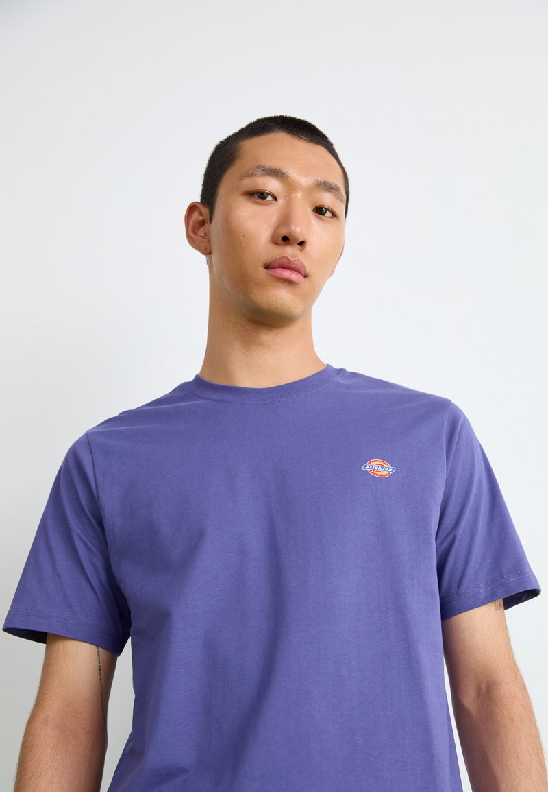 Lila Baumwoll-T-Shirt mit kurzen Ärmeln, Rundhalsausschnitt und einem kleinen bunten Logo auf der Brust. Glatte Textur und lässiges Design.
