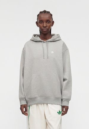 Personne avec des cheveux tressés portant un sweat à capuche gris clair et un pantalon blanc avec des logos verts Adidas, debout devant un fond uni.
