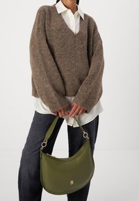 Person klädd i en brun fuzzy tröja över en vit krage skjorta och mörka byxor, håller en olivgrön handväska med guldfärgad hårdvara.