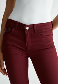 Gros plan d'une personne portant un pantalon moulant rouge foncé avec un bouton argenté et un haut blanc côtelé.