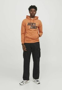 Sudadera naranja con diseño gráfico "JACK & JONES", combinada con pantalones cargo negros y zapatillas blanco-negro, exhibida sobre un fondo neutro.