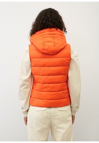 Gepolsterte orange Weste mit Kapuze, ausgestattet mit horizontalen Steppnähten. Über einem cremefarbenen Langarmshirt und hellen Cordhosen getragen.