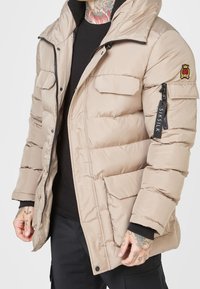 Giacca imbottita beige con chiusura frontale con zip, due tasche frontali e una patch con logo sulla manica. Presenta un colletto testurizzato e dettagli neri.