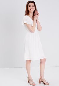 Weisses, knielanges Kleid mit kurzen, flatternden Ärmeln, Knopfdetails und strukturiertem Stoff, kombiniert mit goldenen Absatzsandalen.