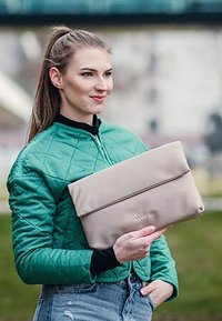 Světlě béžová psaníčka s hladkým povrchem a překlopným designem, drží osoba oblečená v zelené prošívané bundě a džínech.