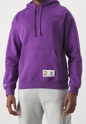Homme portant un sweat à capuche violet des Los Angeles Lakers avec poche avant et écusson du logo NBA, associé à un pantalon de survêtement gris clair.