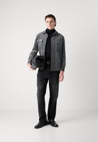 Striped gray jacket with two chest pockets, black turtleneck, black jeans, and black shoes. Holding a black, soft-textured pouch.