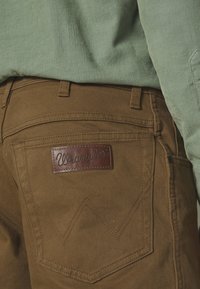 Zbliżenie tylnej kieszeni brązowych jeansów Wrangler z naszywką z logo ze skóry, noszonych z zieloną prążkowaną koszulą.