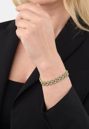 Pulsera en tonos dorado y plateado que presenta eslabones entrelazados con un acabado liso, usada en una mano sobre un fondo de chaqueta oscura.