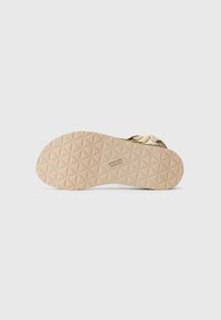 Teva-sandaler med en teksturert beige gummisåle, som har et mønstrete design og beige stropper med en subtil blomsterdetalj.