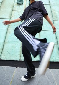 Zwarte oversized T-shirt, donkere trainingsbroek met drie witte strepen en zwarte sneakers met witte zolen. De achtergrond is een groene textuurwand.