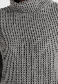 Grauer Rollkragenpullover mit strukturiertem Waffelmuster und geripptem Kragen, der Wärme und eine enge Passform betont.