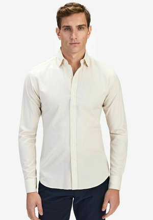 Vaalean beige napillinen paita pitkillä hihoilla ja pehmeällä pinnalla, jossa on klassinen kaulus ja hienovarainen napitus.