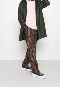 Wodoodporny płaszcz w kolorze oliwkowej zieleni, jasnoróżowa koszulka, brązowe bojówki w kamuflażu oraz czarne trampki z białymi podeszwami i sznurowadłami.