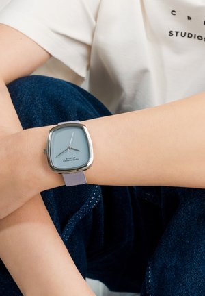 Håndled med et firkantet sølvur med lyseblåt urskive og lavendelfarvet rem, der hviler på krydsede mørkeblå jeans.