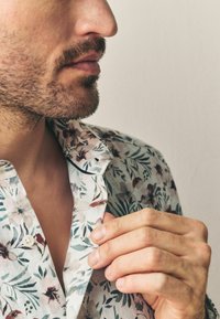 Chemise à boutons à fleurs en tissu léger, présentant une base blanche avec des feuilles vertes et des fleurs roses ; col avec un bord contrastant. Ajustement du col à la main.