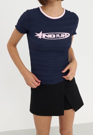 Femme portant un t-shirt bleu marine texturé avec une bordure rose et le texte "NO PLANS", associé à une jupe noire asymétrique.