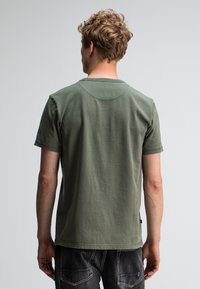 Jonge man met krullend haar, gekleed in een olijfgroen T-shirt met korte mouwen en zwarte spijkerbroeken, met zijn rug naar de camera gekeerd tegen een effen lichte achtergrond.