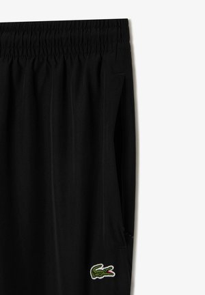 Schwarze sportliche Shorts mit elastischem Bund, Seitentaschen und einem grünen gestickten Krokodil-Logo auf der unteren linken Seite. Glatte Stofftextur.