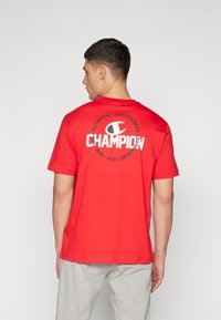 Rød bomulds t-shirt med korte ærmer, der har et stort hvidt "Champion" logo print på ryggen og branded tekst i et cirkulært design.