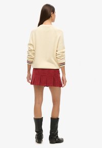 Cremefarvet strikket sweater med ribbede manchetter, parret med en rød fløjl mini nederdel med plisseringer og sorte knæhøje støvler. Enkle, strukturerede designelementer synlige.