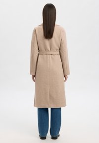 Kvinne med langt, rett hår iført en beige belted frakk, blå jeans og svarte sko, som står vendt bort fra kameraet mot en ensfarget bakgrunn.