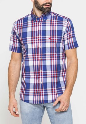 Uomo che indossa una camicia a quadri blu, rossa e bianca a maniche corte con jeans azzurri su sfondo bianco neutro.