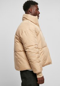 Beige puffjakke med høy krage, glidelås og quiltet tekstur. Har elastiske mansjetter og en løs passform.