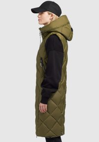 Frau trägt eine lange, gequiltete olivgrüne Kapuzenweste über einem schwarzen Sweatshirt und einer schwarzen Mütze, steht im Profil vor einem hellen Hintergrund.