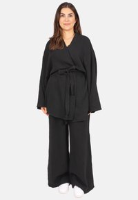 Schwarzes, strukturiertes, leichtes Stoffoutfit mit einer locker sitzenden, kimonoartigen Oberteile und weitem Beinkleid, ergänzt durch weiße Sneakers.