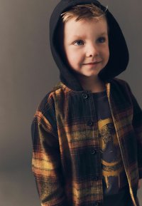 Nupuga särk ruudulise mustriga oranžis, mustas ja kollases. Omab musta kapuutsi ja kantakse halli t-särgi peal, millel on graafiline kujundus.