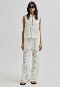 Weißes gesticktes Zweiteiler-Outfit mit einem ärmellosen Top, das eine Bindeschließe hat, und lockeren, langen Hosen mit filigranen Ausschnittmustern.