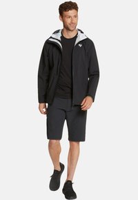 Chaqueta impermeable negra con capucha gris claro, combinada con pantalones cortos negros y zapatillas negras. Presenta un cierre frontal con cremallera y bolsillos.