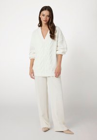 Witte kabelgebreide trui met een V-hals en brede mouwen, gecombineerd met witte wijde broek. Glad textuur en gezellige uitstraling.