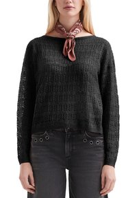 Vrouw in een zwarte gebreide trui met lange mouwen, donkere spijkerbroek met metalen oogjes, en een geknoopt bruine sjaal met patroon.