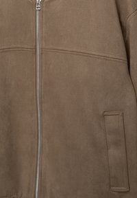 Veste en daim marron avec fermeture éclair argentée verticale, couture horizontale traversant la poitrine, et poche rectangulaire sur le côté inférieur droit.