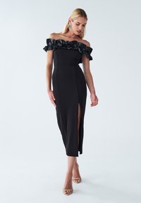 Black off-the-shoulder dress with ruffled neckline, fitted bodice, and thigh-high slit. Smooth fabric with a sleek, elegant silhouette.