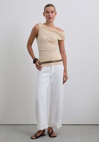 Top asimétrico beige, pantalones de pierna ancha blancos, cinturón negro con tachuelas plateadas, sandalias negras. Tejido suave, corte relajado, diseño casual.