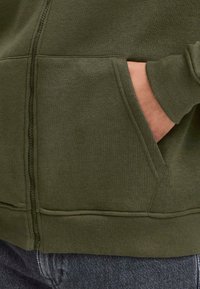 Close-up van een hand in de zak van een olijfgroene hoodie, met de zachte stof en casual stijl tegen donkere spijkerbroeken.