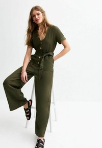 Olive green jumpsuit with short sleeves, V-neck, front buttons, and tie waist. Features wide leg pants and textured fabric. Black sandals.