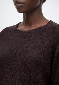 Gros plan d’une personne portant un pull en maille texturée marron foncé avec un col rond, montrant le bas du visage et le haut du torse.